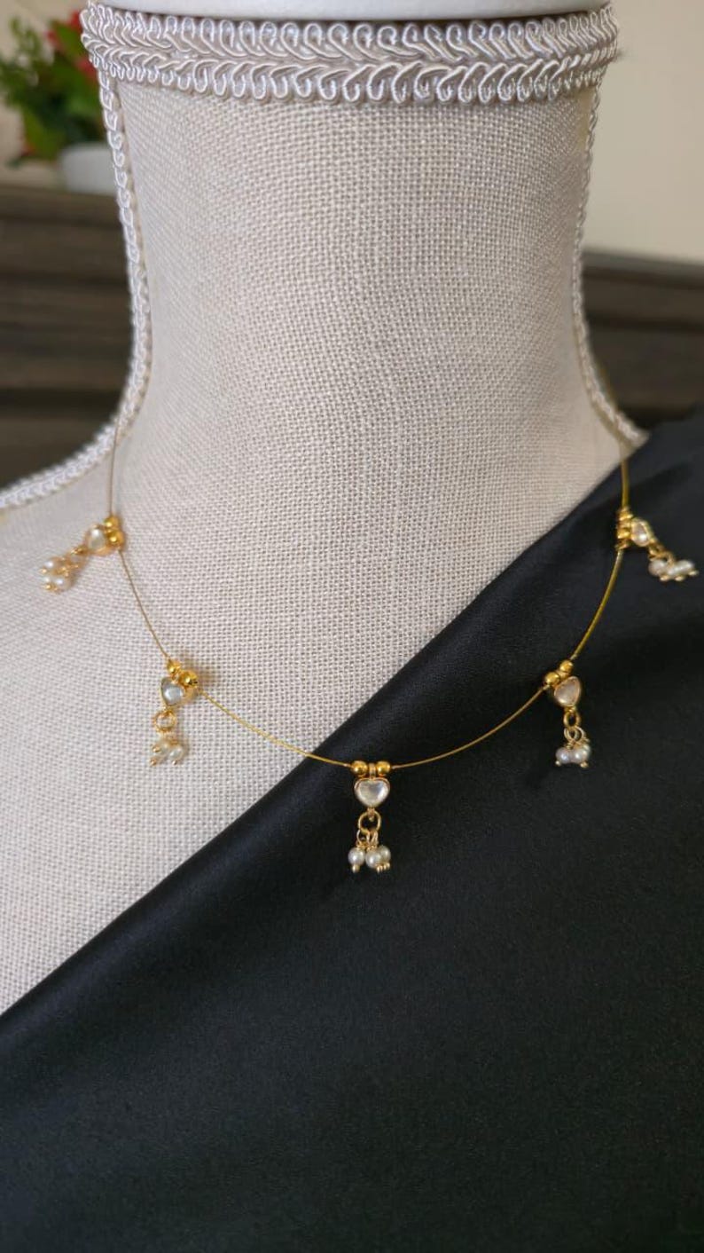 Puede incluir: Delicado collar dorado con colgantes en forma de coraz&oacute;n y peque&ntilde;os detalles de perlas. El collar se exhibe sobre un maniqu&iacute;, con un fondo de tela negra. El collar tiene un dise&ntilde;o minimalista, con una cadena fina y m&uacute;ltiples elementos decorativos.