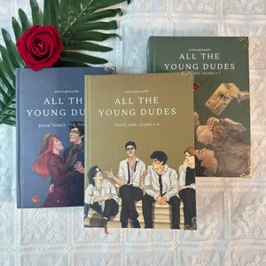May include: Three books titled "ALL THE YOUNG DUDES" are shown, each with unique cover art. The covers feature illustrations and are in shades of blue, green, and tan. A red rose and green foliage are arranged near the books, creating a decorative display.