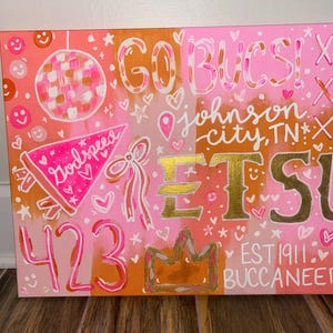 Könnte beinhalten: Eine rosa und orangefarbene Leinwand mit den Worten "GO BUCS!" und "ETSU" in goldfarbenen Buchstaben. Die Leinwand enthält auch die Wörter "Johnson City, TN", "Godspeed", "423" und "EST 1911 Buccaneers".