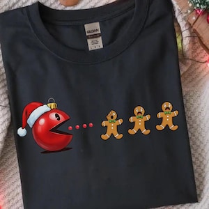Könnte beinhalten: Dunkelgraues T-Shirt mit festlichem Design. Es zeigt einen roten Pac-Man-Charakter mit Weihnachtsmütze, der rote Punkte isst und drei Lebkuchenmänner mit grünen Fliegen jagt. Das T-Shirt besteht aus einem weichen Material.