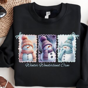 May include: Black sweatshirt featuring three watercolor snowmen in hats and scarves. Each snowman is in a different color scheme: pink, purple, and blue. The text "Winter Wonderland Crew" is below the snowmen.