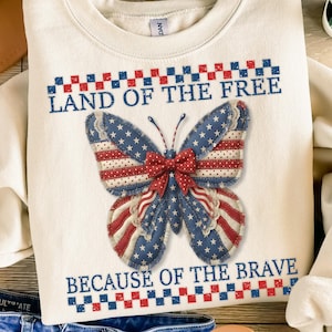 May include: A cream-coloured sweatshirt with a patriotic butterfly design. The butterfly has blue wings with white stars and red and white striped accents. The text "LAND OF THE FREE" and "BECAUSE OF THE BRAVE" are printed above and below the butterfly.