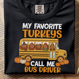 Op de afbeelding: Zwart T-shirt met de tekst "MY FAVORITE TURKEYS CALL ME BUS DRIVER". Het ontwerp toont een gele schoolbus met cartoon kalkoenen erin en pompoenen eromheen. Het shirt heeft een Comfort Colors label.