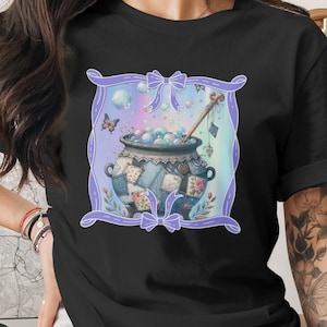 以下が含まれることがあります： 泡と木製のかき混ぜスプーンが入った大釜のファンタジーイラストが描かれた黒いTシャツ。大釜はパッチワーク生地で装飾され、リボンと蝶が付いたラベンダー色のフレームで囲まれています。