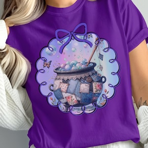 Op de afbeelding: Paarse t-shirt met een grillige illustratie van een ketel gevuld met bellen, omgeven door een geschulpte lijst en een strik. De ketel heeft een patchwork-ontwerp met bloemen- en patroonpatches.
