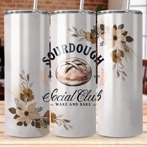 May include: White stainless steel tumbler with a brown sourdough bread design and floral accents. The tumbler has the text "Sourdough Social Club - Wake and Bake" printed on it.