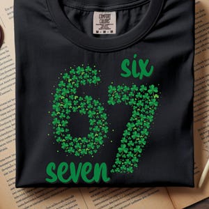Puede incluir: Camiseta negra con un diseño de tréboles verdes que forman los números "67" y las palabras "six" y "seven". La camiseta tiene cuello redondo y es de material suave.