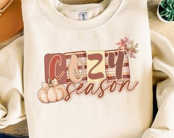 May include: Cream-colored sweatshirt with the words "COZY season" in a fall-themed design. The design includes a pumpkin, plaid pattern, and autumn leaves. The sweatshirt is made of a soft material.