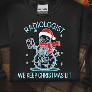 Könnte beinhalten: Schwarzes Sweatshirt mit einem Schneemann-Design, der einen Weihnachtsmannhut und einen Schal trägt, mit einem Skelett im Inneren. Der Text lautet "RADIOLOGIST" darüber und "WE KEEP CHRISTMAS LIT" unter der Schneemann-Grafik.
