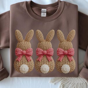 Puede incluir: Sudadera marrón con tres diseños de conejitos de punto. Cada conejito tiene un lazo rosa y una cola de pompón blanco. La sudadera tiene cuello redondo y mangas largas, ideal para Pascua.