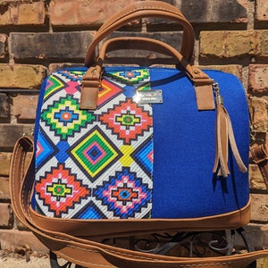 May include: A blue and multi-colored geometric patterned handbag with a brown leather strap and handle. The bag has a small brown tassel hanging from the handle.