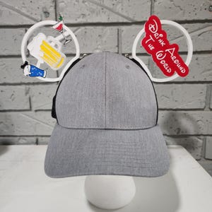Puede incluir: Gorra de béisbol gris con la parte trasera de malla negra. La gorra tiene detalles de orejas de Mickey Mouse blancas con gráficos temáticos de bebidas y el texto "Drink Around The World" en rojo.