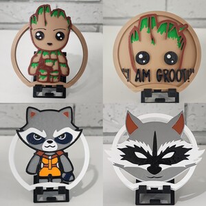 Puede incluir: Cuatro soportes para teléfono impresos en 3D con personajes de la película Guardianes de la Galaxia. Dos soportes representan a Groot, uno con un diseño verde y marrón y el otro con el texto "I AM GROOT!" Dos soportes representan a Rocket Raccoon, uno con un diseño gris y negro y el otro con un diseño gris y blanco.