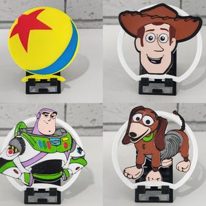 Puede incluir: Cuatro figuras de personajes de Toy Story en soportes blancos. Los personajes son Woody, Buzz Lightyear, Slinky Dog y la pelota de la película. Las figuras son de plástico y están pintadas en colores brillantes.