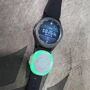 Puede incluir: Un reloj inteligente negro con un icono de Mickey Mouse en la banda. La esfera del reloj muestra la hora, la fecha y la temperatura. El reloj est&aacute; sobre una superficie gris.
