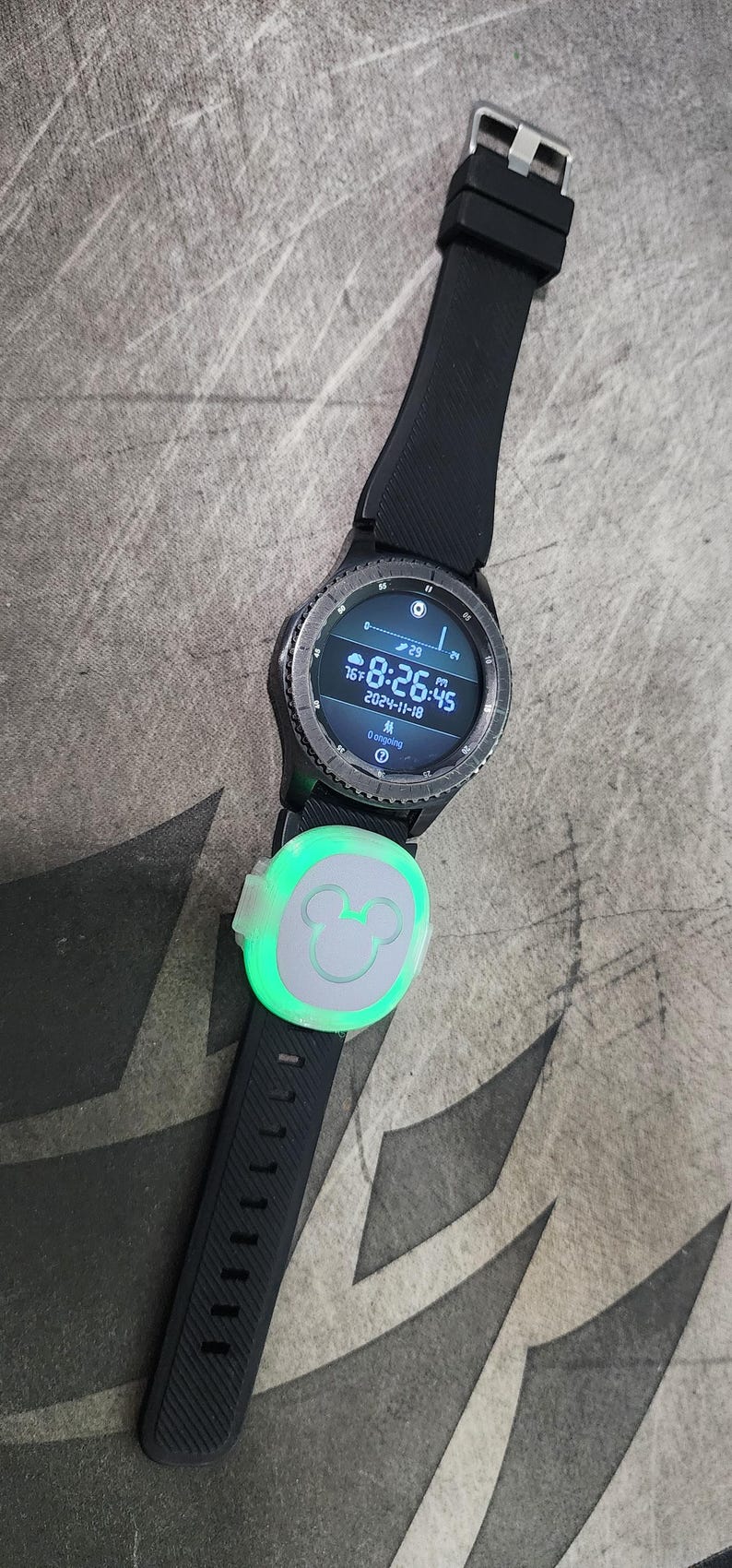 Puede incluir: Un reloj inteligente negro con un icono de Mickey Mouse en la banda. La esfera del reloj muestra la hora, la fecha y la temperatura. El reloj est&aacute; sobre una superficie gris.
