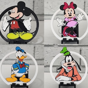 May include: Four Disney character figurines: Mickey Mouse, Minnie Mouse, Donald Duck, and Goofy. Each character is set within a white circular frame and mounted on a black base. Mickey is in black, red, and yellow. Minnie is in pink and red. Donald is in blue and yellow. Goofy is in orange and green.
