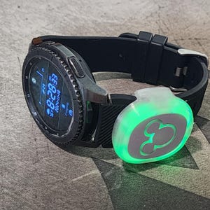 May include: A black smartwatch with a green Mickey Mouse light-up accessory attached to the band. The watch face displays the time, date, and heart rate.