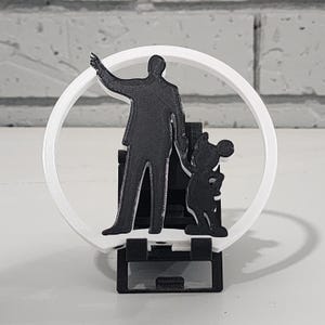 Puede incluir: Escultura en blanco y negro impresa en 3D que presenta la silueta de un hombre con el brazo levantado, junto a la silueta de Mickey Mouse. Las figuras están dentro de un marco circular blanco y montadas sobre una base negra.