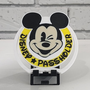 Puede incluir: Un soporte para pases de Disney blanco con la cara sonriente de Mickey Mouse con orejas negras. El círculo amarillo alrededor de Mickey tiene las palabras "DISNEY PASS HOLDER" en negro. El soporte tiene una base negra.