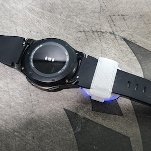 Puede incluir: Un reloj inteligente negro con una esfera redonda y una banda de goma negra. El reloj est&aacute; sobre una superficie gris.