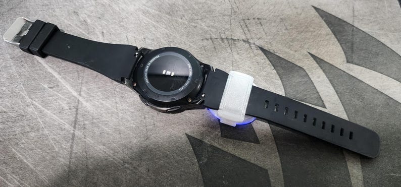 Puede incluir: Un reloj inteligente negro con una esfera redonda y una banda de goma negra. El reloj est&aacute; sobre una superficie gris.