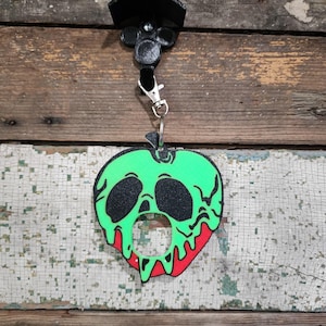 Puede incluir: Un llavero en forma de manzana verde y negra con un diseño de goteo rojo. El llavero está sujeto a un clip negro y a un soporte negro. El diseño de la manzana tiene un aspecto espeluznante y de dibujos animados.