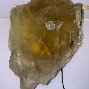 Puede incluir: Un gran cristal de calcita translúcida de color ámbar con una forma irregular y rugosa. Una moneda se encuentra en la superficie para comparar el tamaño. El cristal tiene un aspecto natural, sin pulir, mostrando su estructura cristalina.