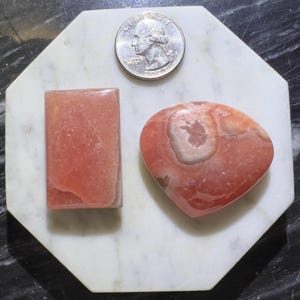 May include: Two pink and white gemstone cabochons, one rectangular and one heart-shaped, on a white marble surface.