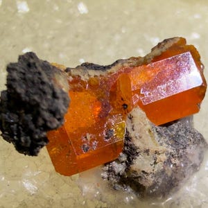 May include: A vibrant orange wulfenite crystal specimen featuring a translucent, tabular structure with sharp, geometric edges. The crystal is attached to a dark, textured, greyish-brown mineral matrix, showcasing a contrast between the smooth, glassy orange surface and the rough, earthy base material.