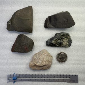 May include: Five irregularly shaped rocks of varying colors and textures are displayed on a white surface. A quarter is next to one of the rocks for size comparison. A ruler is at the bottom of the image.