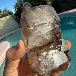 Puede incluir: Un gran cristal de cuarzo ahumado en bruto sostenido en una mano. El cristal es de color marrón translúcido con estructuras internas y facetas visibles. El fondo incluye una piscina y vegetación.
