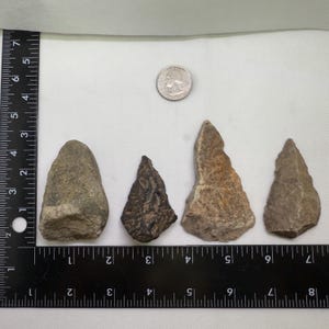 Puede incluir: Cuatro piedras de forma irregular de varios tamaños y colores, que van del gris al marrón, están dispuestas sobre una superficie blanca. Se coloca una moneda cerca de las piedras para comparar el tamaño. Una regla se coloca junto a las piedras.