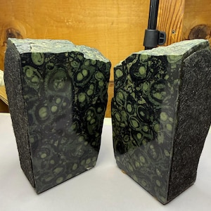 May include: Two bookends made of polished stone. The stone has a dark background with green and light green circular patterns. The sides of the bookends are covered in a dark, textured material. The bookends are rectangular with uneven tops.