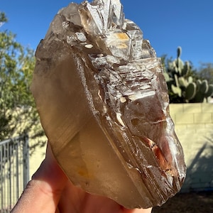 May include: A large, raw smoky quartz crystal with a translucent, amber-brown hue. The crystal has a rough, natural texture with visible internal fractures and inclusions. The background features a clear blue sky and desert vegetation.