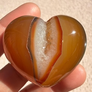 Könnte beinhalten: Ein polierter, herzförmiger Achatstein. Der Stein hat eine warme, bernsteinfarbene Außenseite mit einem weißen und orange-roten Inneren. Das Herz wird in einer Hand gehalten, was seine glatte, glänzende Oberfläche und natürlichen Muster zeigt.