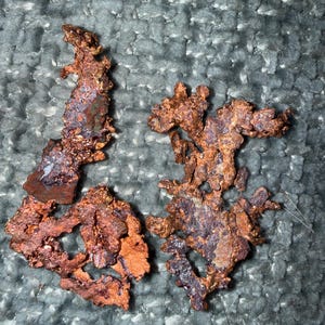 May include: Two raw, unrefined copper specimens with irregular, jagged shapes and a metallic, oxidised surface texture. The pieces exhibit a mix of bright copper-orange and dark, weathered brown tones, showcasing a natural, porous, and crystalline structure.