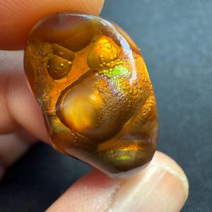 Könnte beinhalten: Ein polierter, oval geformter Edelstein mit einer warmen Farbpalette. Der Stein weist eine Mischung aus Bernstein-, Braun- und Goldtönen mit schillernden grünen und orangen Bereichen auf. Die Oberfläche hat ein strukturiertes, blasenartiges Aussehen. Der Stein wird zwischen zwei Fingern gehalten.