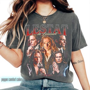 Op de afbeelding: Donkergrijs t-shirt met een vintage design. De afbeelding toont de woorden "LESTAT de Lioncourt" in rood en wit, en meerdere portretten van een persoon. Het shirt heeft een distressed look, met "pepper comfort colors" onderaan gedrukt.