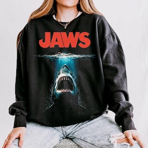 May include: Black sweatshirt featuring the word "JAWS" in red block letters above an image of a shark emerging from the ocean. The shark's mouth is open, revealing sharp teeth. The design is a vintage movie-themed graphic.