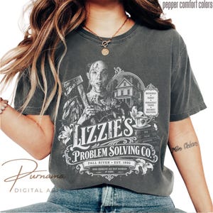 Puede incluir: Una camiseta de manga corta gris carbón con un estampado gráfico blanco de una persona sosteniendo un hacha, una casa y una taza de té. El texto dice Lizzie's Problem Solving Co., Fall River, Est. 1892 y enumera household family persistent issues.