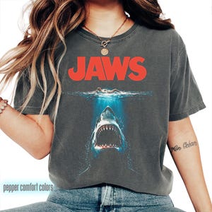 Op de afbeelding: Donkergrijs T-shirt met het woord "JAWS" in het rood boven een haaienillustratie. De afbeelding toont een zwemmer in het water. Het shirt heeft een vintage look en is gemaakt van zacht materiaal.