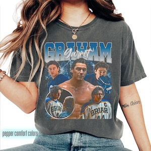 May include: A charcoal gray short-sleeve t-shirt featuring a graphic print with the text GRAHAM and Garret. The design includes multiple photographic images of a hockey player in a Briar jersey and a shirtless portrait on a blue background.