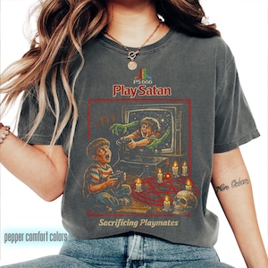 Puede incluir: Camiseta gris oscuro con un gráfico de estilo vintage. El diseño presenta el texto "Play Satan" sobre una ilustración de un niño jugando a un videojuego, con una mano demoníaca extendiéndose. La frase "Sacrificing Playmates" está debajo.