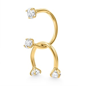 May include: A gold-coloured, double-sided piercing ring with four clear, round-cut gemstones. The ring is knotted in the centre, with the gemstones at each end of the curved design. The jewellery is set against a white background.
