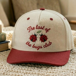 May include: A baseball cap with a beige crown and a maroon brim. The cap features embroidered ladybugs and the text "The last of the bugs club" in maroon thread. A small maroon button is on top.