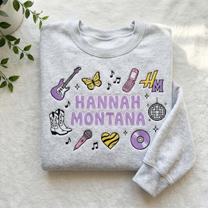 May include: A light gray sweatshirt with the text "HANNAH MONTANA" in purple. The sweatshirt features various embroidered patches, including a guitar, butterfly, phone, cowboy boots, microphone, heart, and a disco ball.