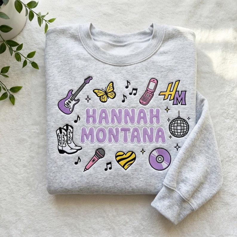 May include: A light gray sweatshirt with the text "HANNAH MONTANA" in purple. The sweatshirt features various embroidered patches, including a guitar, butterfly, phone, cowboy boots, microphone, heart, and a disco ball.