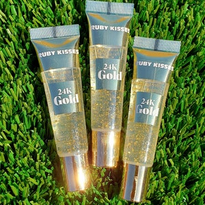 May include: Three clear lip gloss tubes with gold accents and gold flakes inside, labeled "RUBY KISSES" and "24K Gold". The tubes are arranged on a bed of green artificial grass, creating a luxurious and cosmetic-focused aesthetic.
