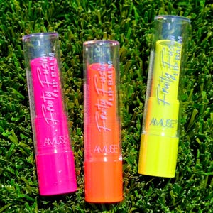 May include: Three tubes of lip balm in vibrant neon colors: pink, orange, and yellow. Each tube is labeled "Fruity Fusion Lip Balm" and "AMUSE" in white text. The lip balms are displayed on a bed of green artificial grass.